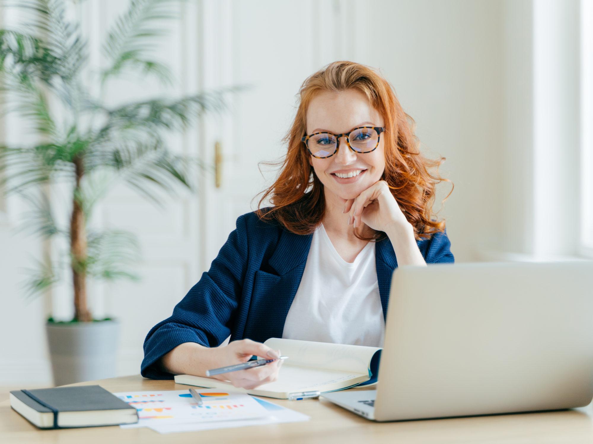 Foto von erfolgreicher Freiberuflerin im HomeOffice, schaut Webinar online auf Laptop, schreibt Informationen in Notizblock, lächelt fröhlich, posiert am Arbeitsplatz, in formeller Kleidung gekleidet
