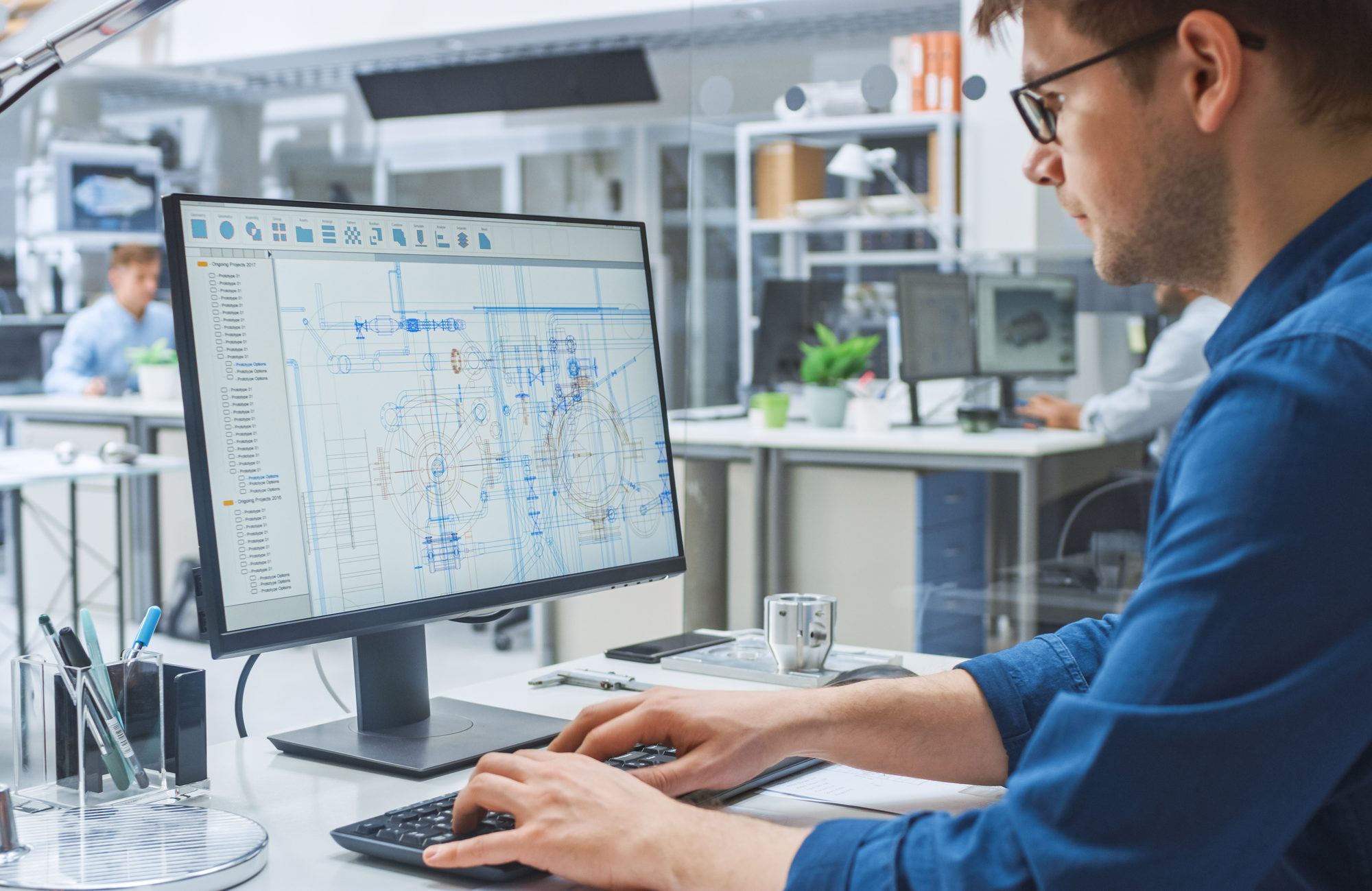Blick über die Schulter eines Technikers, der eine CAD-Software auf einem Desktop verwendet, auf dem Bildschirm sind technische Entwürfe und Zeichnungen zu sehen. Im Hintergrund Fertigungsanlage für Industriedesign.