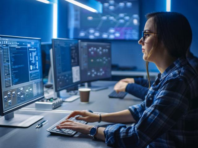 Intelligente junge IT-Programmiererin arbeitet am Desktop PC, grüner Bildschirm, im Kontrollraum des Datencenters. Team von jungen Angestellten programmiert anspruchsvollen Code